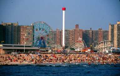 Crowded beach scene with blue water, amusement park rides including a Ferris wheel and tall tower, and city buildings in the background