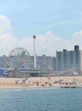 A beachside amusement park with a Ferris wheel, tall rides, and city buildings in the background under a bright blue sky.