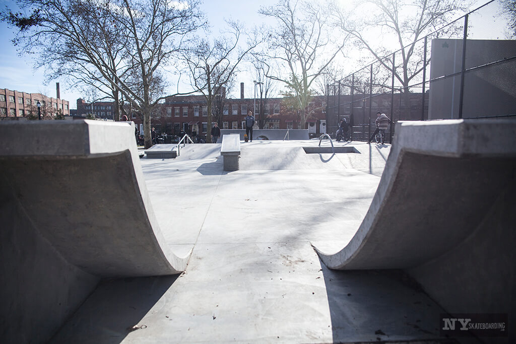 The image shows a concrete skate park with ramps and rails, framed by bare trees and surrounded by a metal fence on a sunny day.
