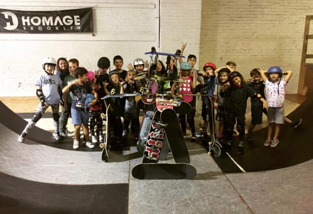 The image shows a group of children wearing helmets and protective gear, posing with skateboards and scooters on an indoor skate ramp at Homage Skateboard Academy.