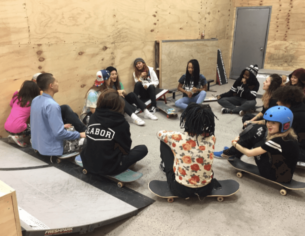 The image shows a group of young people sitting in a circle indoors, most sitting on skateboards, engaged in conversation at a skate school or community center.