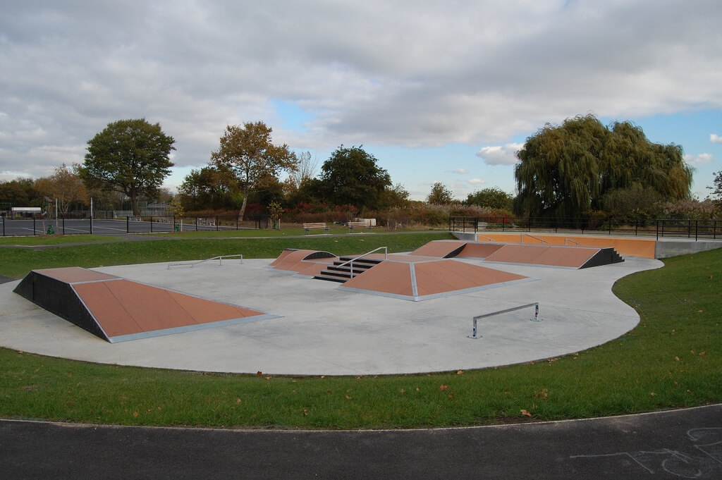 The image shows a small concrete skatepark with multiple ramps and rails, surrounded by grass and trees under a cloudy sky.