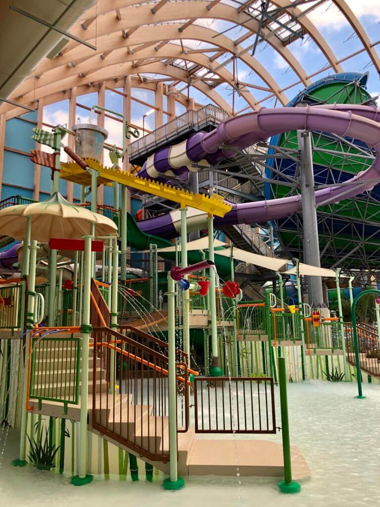
Indoor water park features colorful slides, climbing structures, water sprayers, and play areas under a large glass dome ceiling. The image shows an indoor water park with colorful slides, climbing structures, and water play features under a large glass roof.