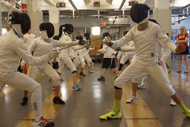 two rows of fencers stand across from each other and compete with foil swords