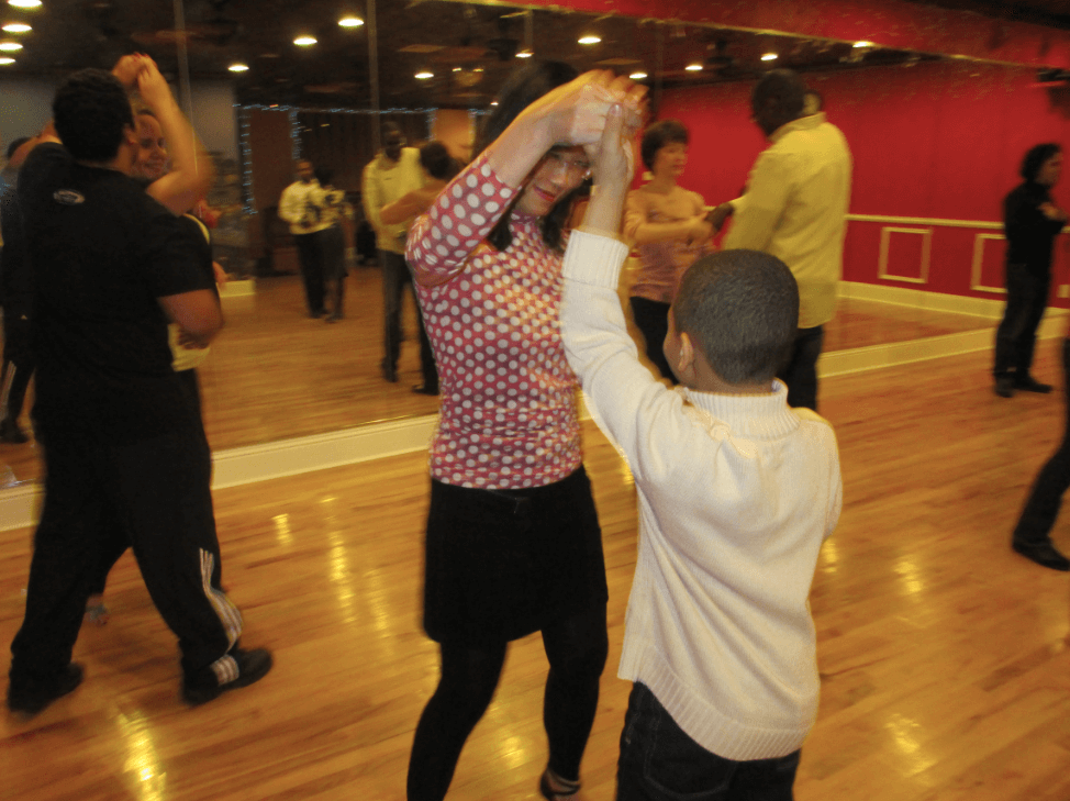 People participate in a dance class in a studio with mirrored walls; a woman and a child hold hands and dance on a polished wooden floor.