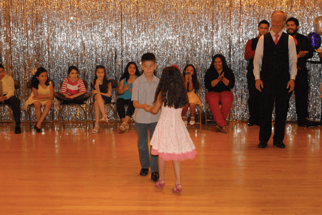 
The image "mende-dance-studio.jpg" shows two children dancing together in the middle of a studio with a shiny silver backdrop, while people watch from chairs along the wall. An adult stands to the right, observing the scene.