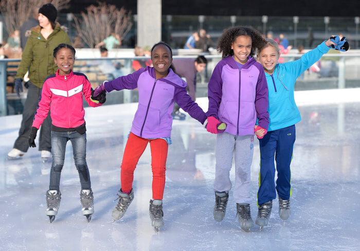Four smiling children ice skating outdoors on a sunny winter day, dressed in colorful jackets and holding hands as they enjoy the activity together.