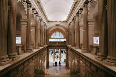 Grand interior hall of a museum with tall classical columns, arched ceiling, and visitors viewing artwork displays on two levels.