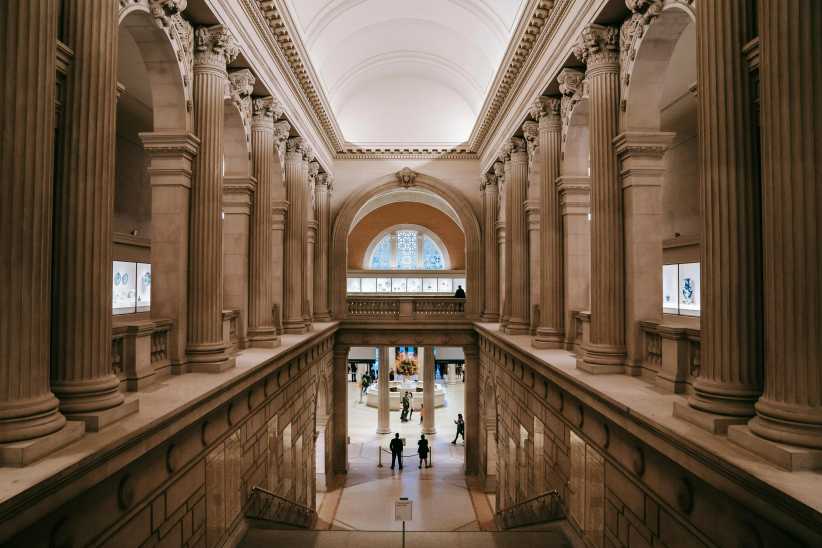 Grand interior hall of a museum with tall classical columns, arched ceiling, and visitors viewing artwork displays on two levels.
