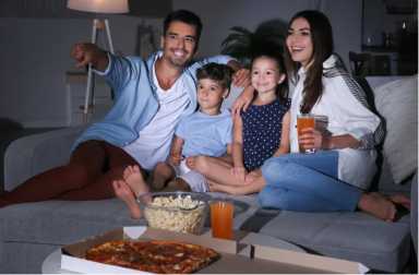A family of four enjoys watching television; the father points at the screen while the mother holds drinks. They are sitting on a couch in a dimly lit living room, surrounded by pizza and popcorn.
