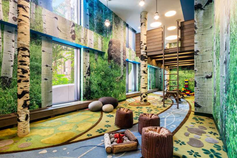 The image shows a playroom with birch tree decor, soft seating, large windows, colorful rugs, and wooden climbing structures.The image displays a nature-themed children's playroom with birch tree decor, soft seats, climbing equipment, and colorful blocks.