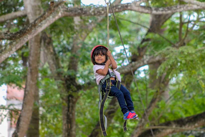 The kid zip‑lining over trees, wearing helmet and safety harness, with green forest underneath.