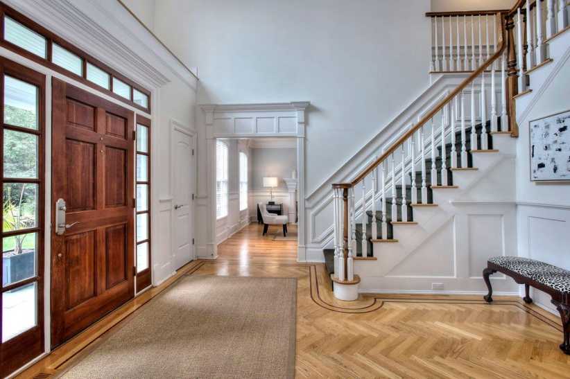 
A spacious entryway features a wooden front door, hardwood floors, white walls, a staircase with white spindles, and natural light.