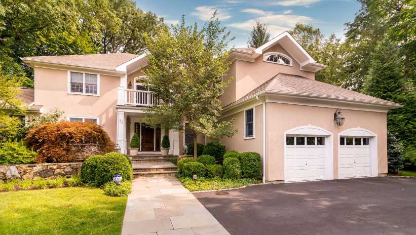 A two-story beige house with white trim, a double garage, and manicured landscaping sits surrounded by lush trees and greenery.