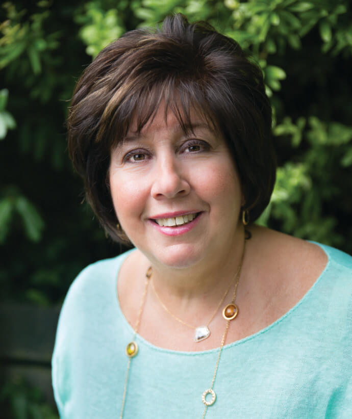 A woman with short brown hair wears a light blue top and gold necklaces, standing outdoors in front of green leafy bushes. A woman with short brown hair wears a light blue top and gold necklaces, standing outdoors in front of green leafy bushes.