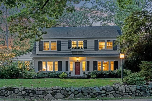 The image shows a two-story house with gray siding, black shutters, and a red front door, warmly lit from inside, with a stone wall and lush landscaping in front.