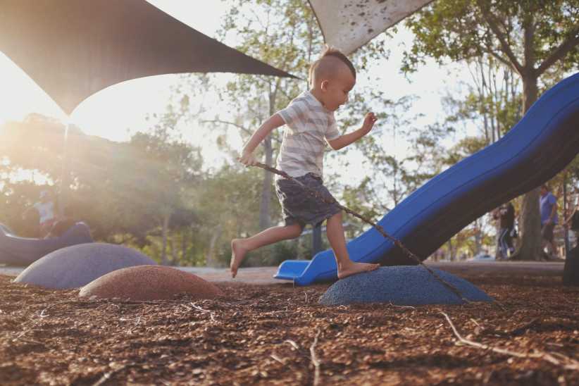 A young child runs barefoot across soft playground mounds, holding a stick with a blue slide and trees in the sunny background.