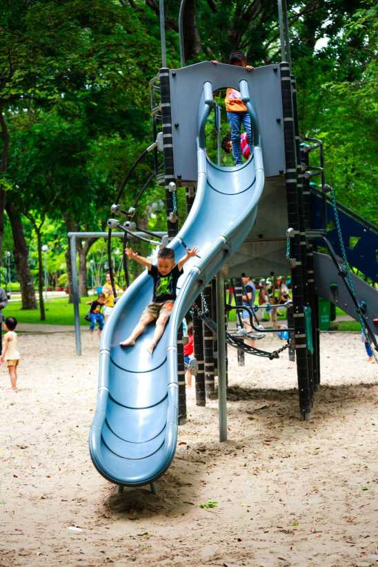 Children play on a tall playground structure with a curved slide, landing in soft sand while others climb and play in the background.