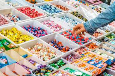 A person selects candy from a display of assorted colorful wrapped candies and chewing gum packs arranged in plastic trays.