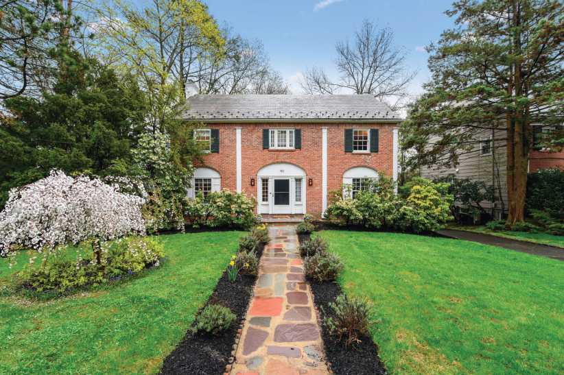 A charming suburban home with peaked roof, wide front porch, stone & siding façade, set amid verdant trees and landscaped bushes.