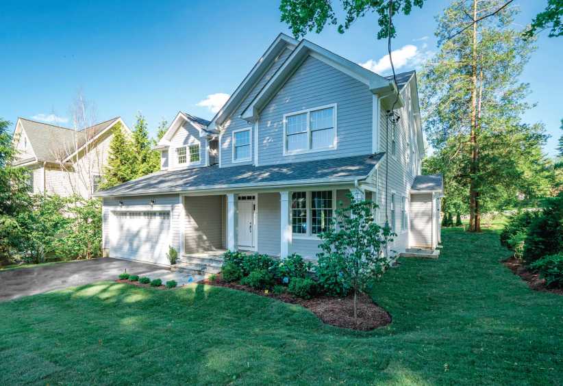 Two-story suburban house with gray siding, white trim, garage, and landscaped yard surrounded by trees on a sunny day.