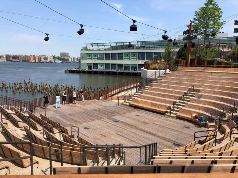 An outdoor wooden amphitheater by the water with tiered seating, overhead lights, and a group of people near the waterfront.