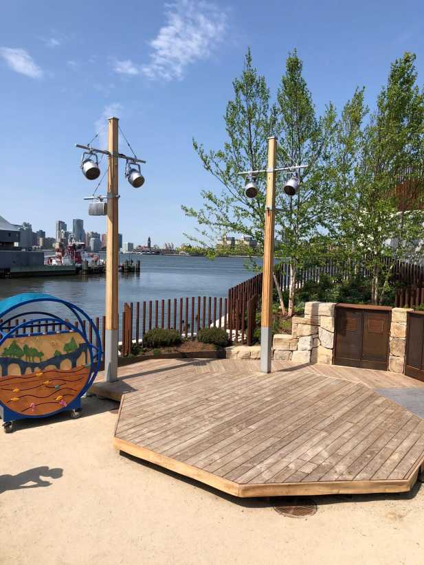 Wooden octagonal stage with lights, near waterfront with trees and city skyline in the background on a sunny day.