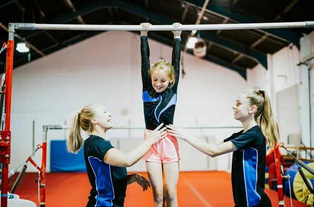 Kids in a gymnastics gym: one little girl hangs from a bar while an instructor or adult stands nearby.