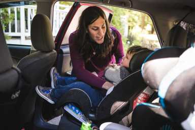 A woman leans into a car to secure a young child in a rear car seat, smiling and talking while ensuring the seatbelt is fastened.