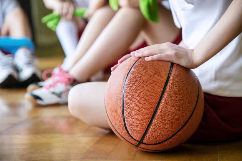 A person sits on a gym floor with a basketball under their hand, while others with athletic shoes and uniforms sit nearby, out of focus.