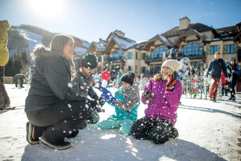 A happy family plays in the snow at a winter resort with ski lodges, dressed in warm clothing, with sunlight shining brightly.