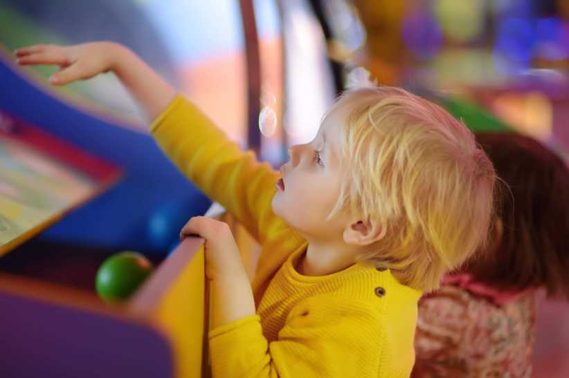 A young child in a yellow sweater reaches out to play an arcade game, with another child nearby in a brightly colored indoor setting.