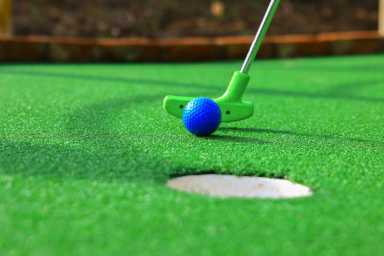 A blue golf ball and green putter are about to strike on bright green artificial turf, aiming for a hole in a mini-golf course.
