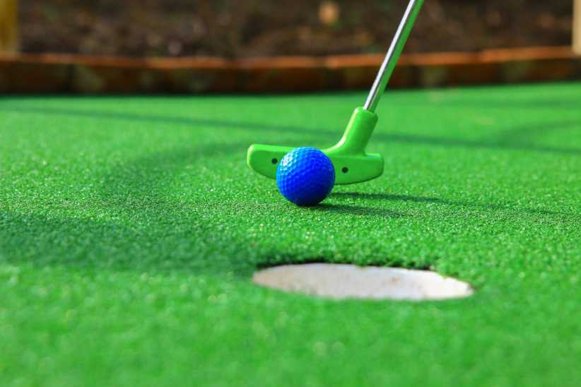 A blue golf ball and green putter are about to strike on bright green artificial turf, aiming for a hole in a mini-golf course.