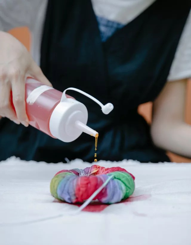 A person squeezes dye from a bottle onto a scrunched, rainbow-colored fabric bundle, starting the tie-dye process on a table.