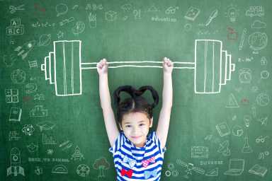 A young girl stands in front of a chalkboard with doodles, raising her arms as if lifting a barbell drawn on the board above her head.