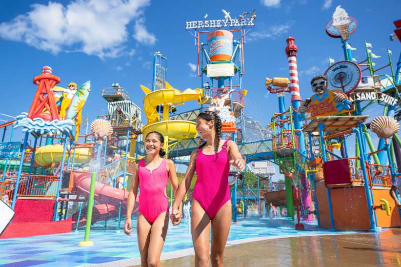 Two girls in matching pink swimsuits walk hand in hand through a colorful water play area at Hersheypark’s Boardwalk.