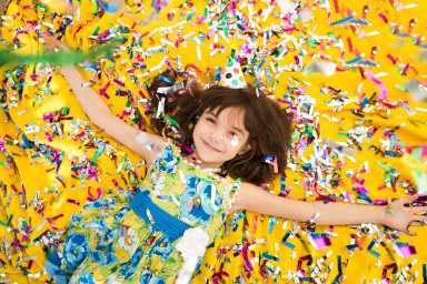 A girl in a blue floral dress and party hat lies on a yellow surface covered in colorful confetti, celebrating joyfully.A girl in a blue floral dress and party hat lies on a yellow background, surrounded by colorful confetti at a festive party.