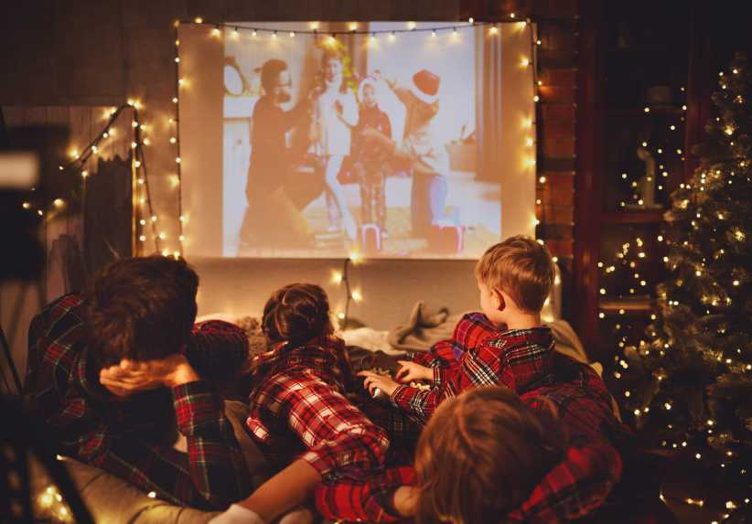 Family in matching plaid pajamas watching a Christmas movie on a projector, surrounded by festive string lights and a decorated tree.