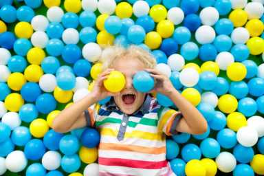 A child in a colorful striped shirt lies on a ball pit filled with blue, yellow, and white plastic balls, playfully holding two balls.