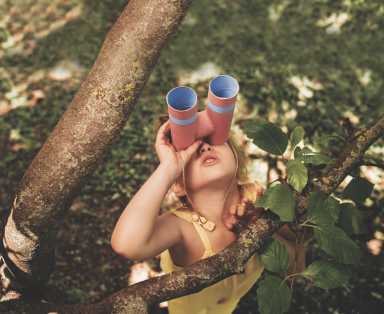 A child in a yellow dress is climbing a tree, surrounded by branches and green leaves, with sunlight filtering through the foliage.
