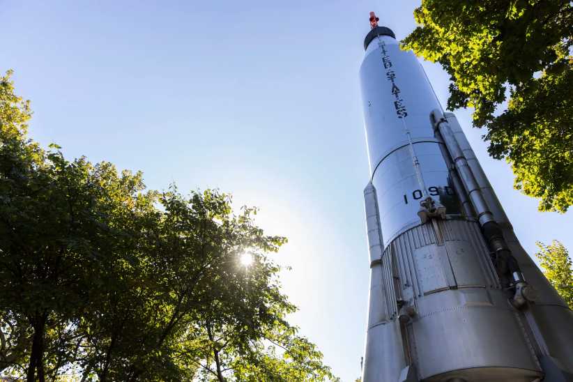 The image shows a tall silver rocket with "United States" written on it, surrounded by trees and sunlight shining through leaves.