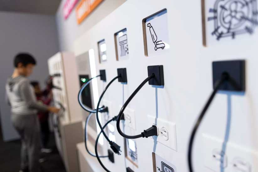 The image shows a hands-on exhibit with power cords plugged into wall sockets, with icons for appliances and people interacting nearby.