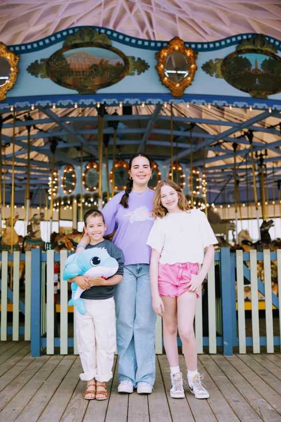 Three children pose in front of a colorful carousel; one holds a blue plush toy, and the others stand smiling on the wooden platform.