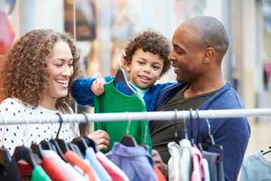 A family with blurred faces shops for clothes together, examining colorful shirts on a rack inside a retail store.