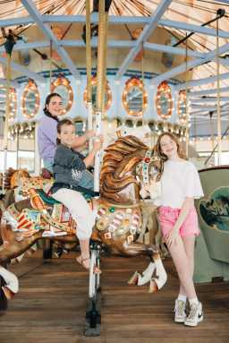Three children enjoy a carousel ride, with two sitting on a decorated horse and one standing beside, in a light-filled amusement park setting.