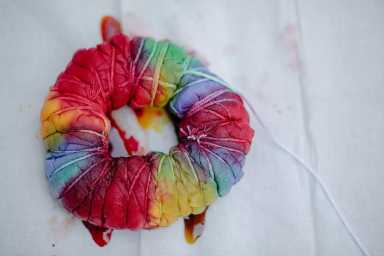 A tightly wrapped fabric bundle with rubber bands, dyed in bright rainbow colors, sits on a white surface for tie-dyeing.