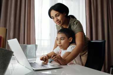 A woman helps a young girl with homework as they sit at a table, using a laptop in a sunlit room with curtains.