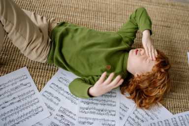 A red-haired child in a green sweater lies on a rug, covering their face, surrounded by scattered sheets of music.