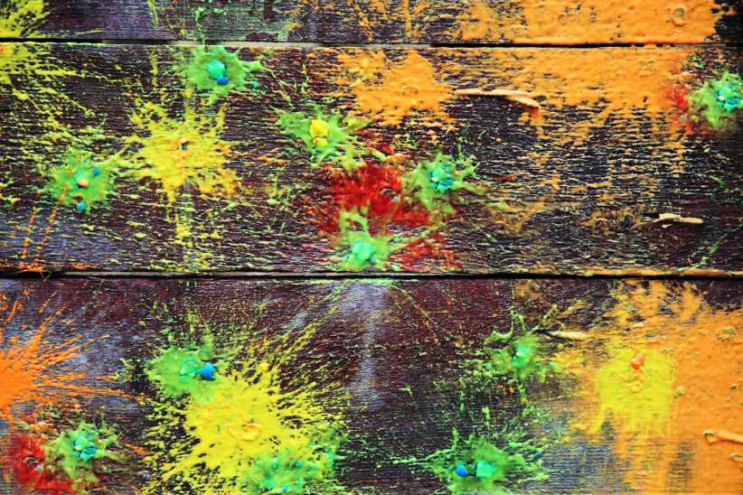 Colorful paintball splatters in yellow, orange, and green cover wooden boards, creating a vibrant, abstract burst effect on the surface.